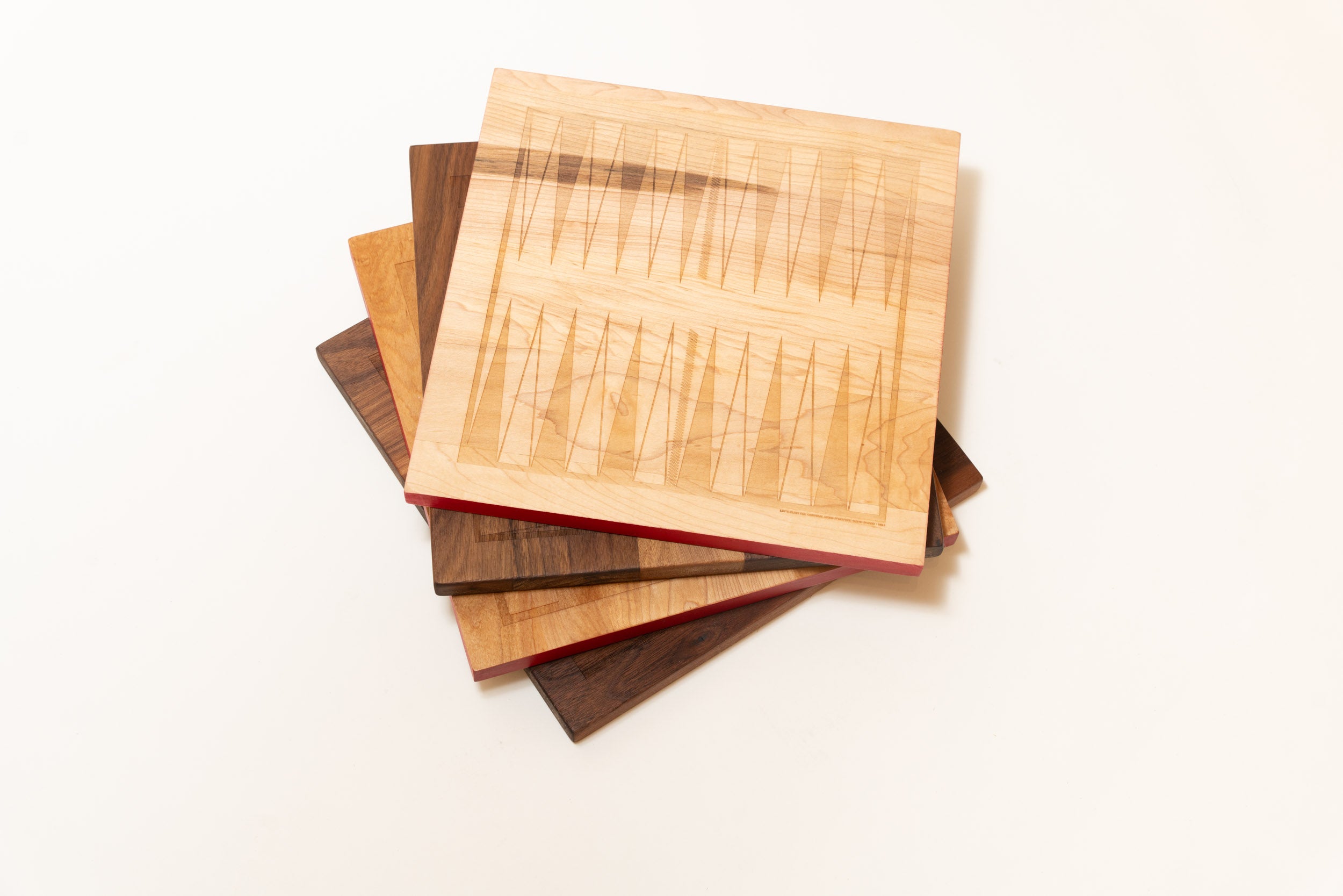 A fanned stack of five Snackgammon boards on a white background, the top one in maple showing the etched backgammon points.