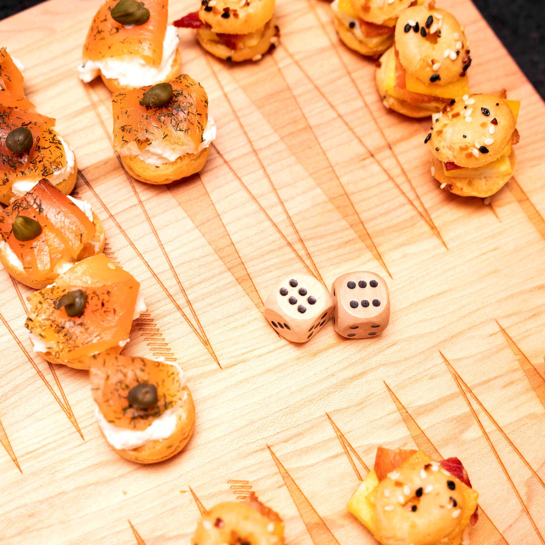 Bagel bites topped with smoked salmon and capers standing in as playing pieces on the etched backgammon points of a North American maple Snackgammon board, two dice at the center.