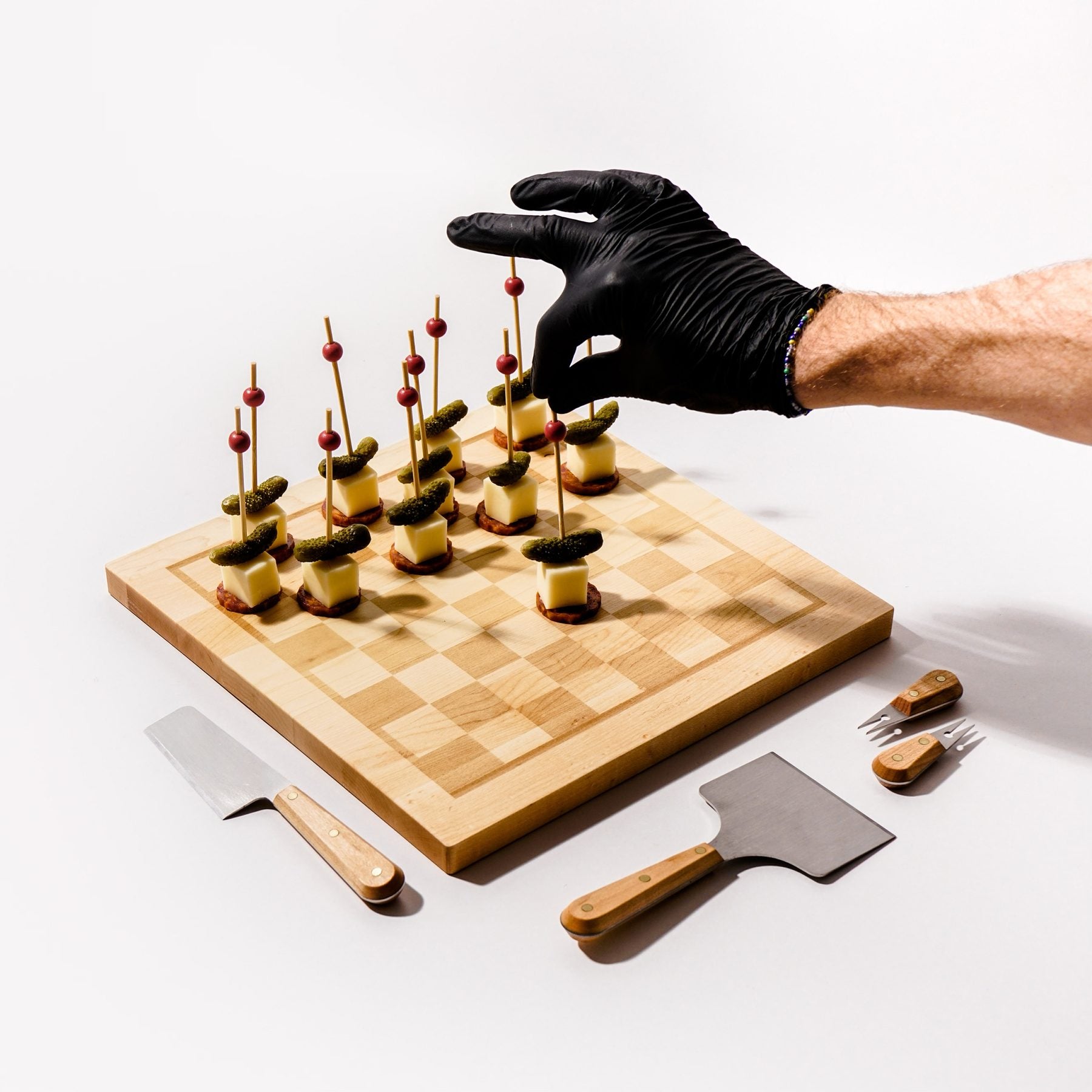 A gloved hand setting a cheese-and-pickle bite onto a maple Snackgammon board etched with the eight-by-eight game grid, cheese knives laid alongside.